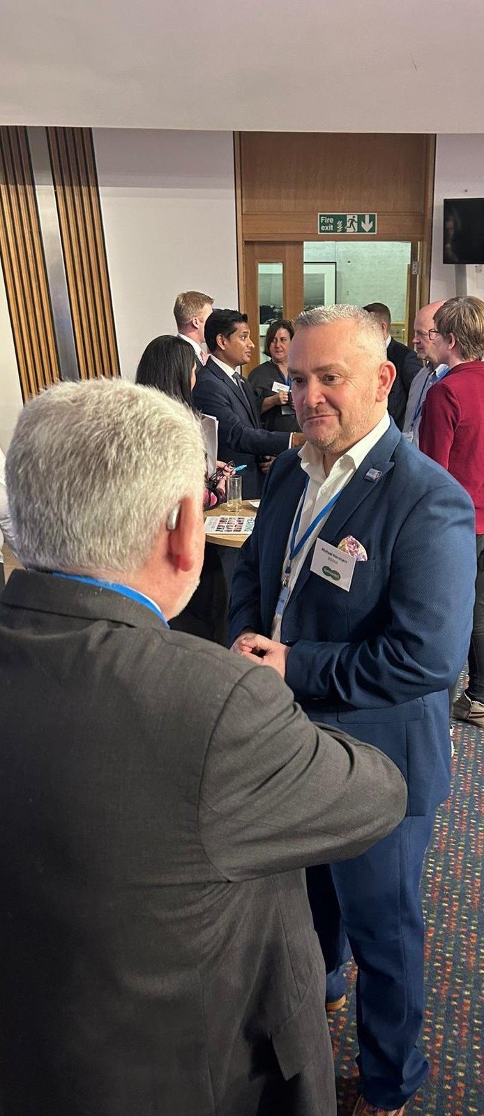 Michael Marchant speaking with attendees inside the Scottish Parliament at Holyrood during a hearing‑health awareness event.