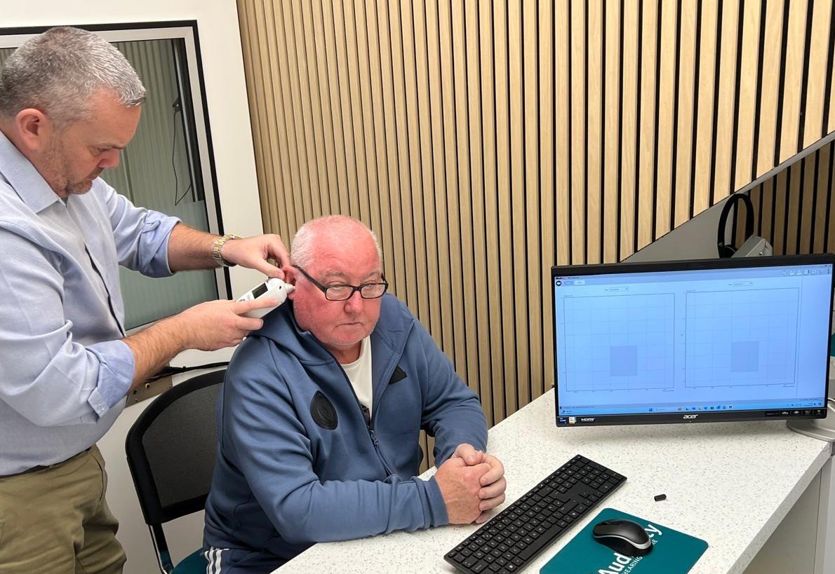 Clinician performing a hearing assessment with a handheld device beside a patient, with audiogram results displayed on a computer screen in a modern hearing clinic.